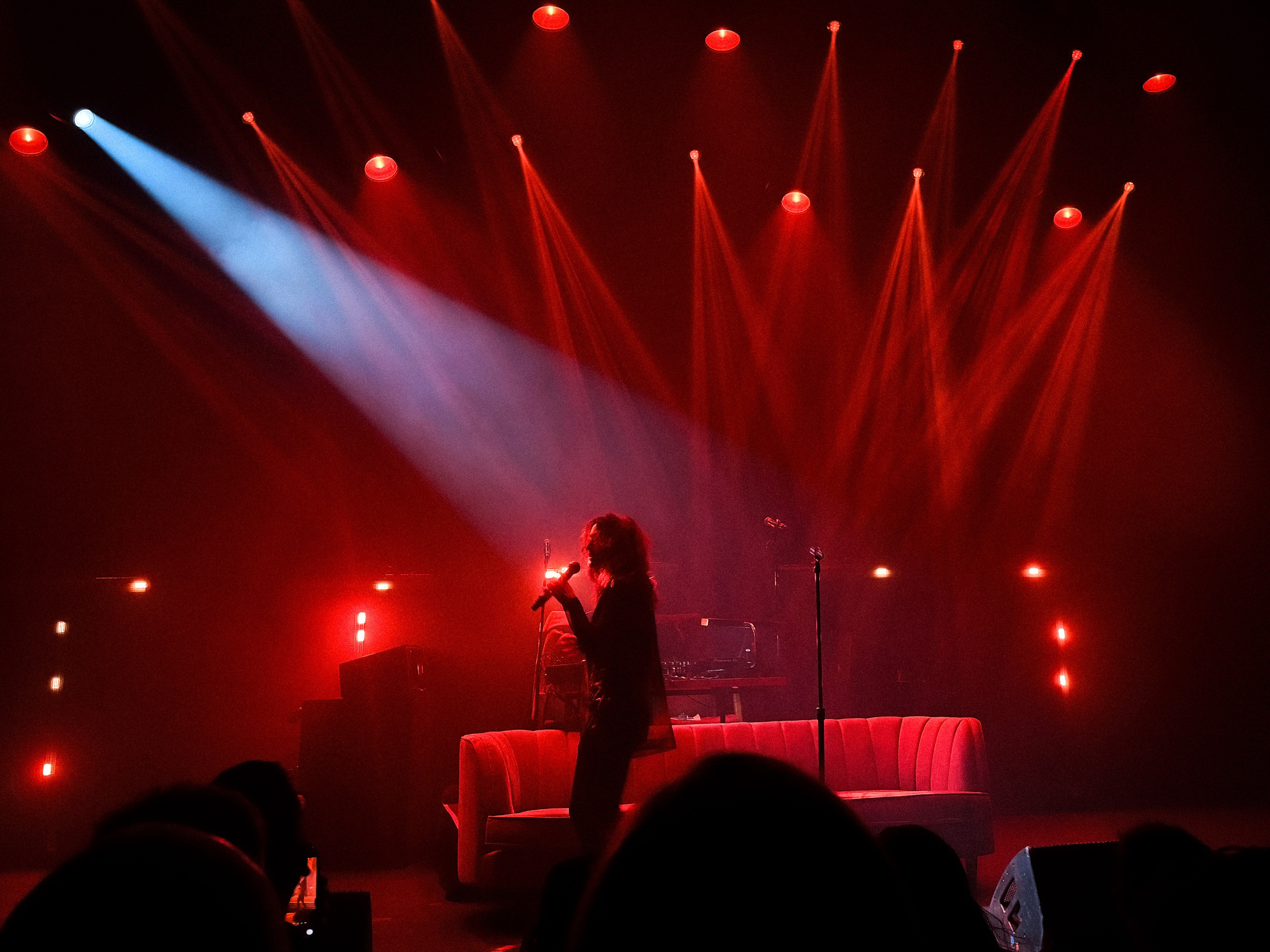 Dragonette sings into a mic on a dim stage drenched in deep red light, with a strong diagonal beam of blue cutting across the scene. She stands near a low white couch, silhouette crisp against the dramatic lighting, while the DJ table glows faintly behind her.