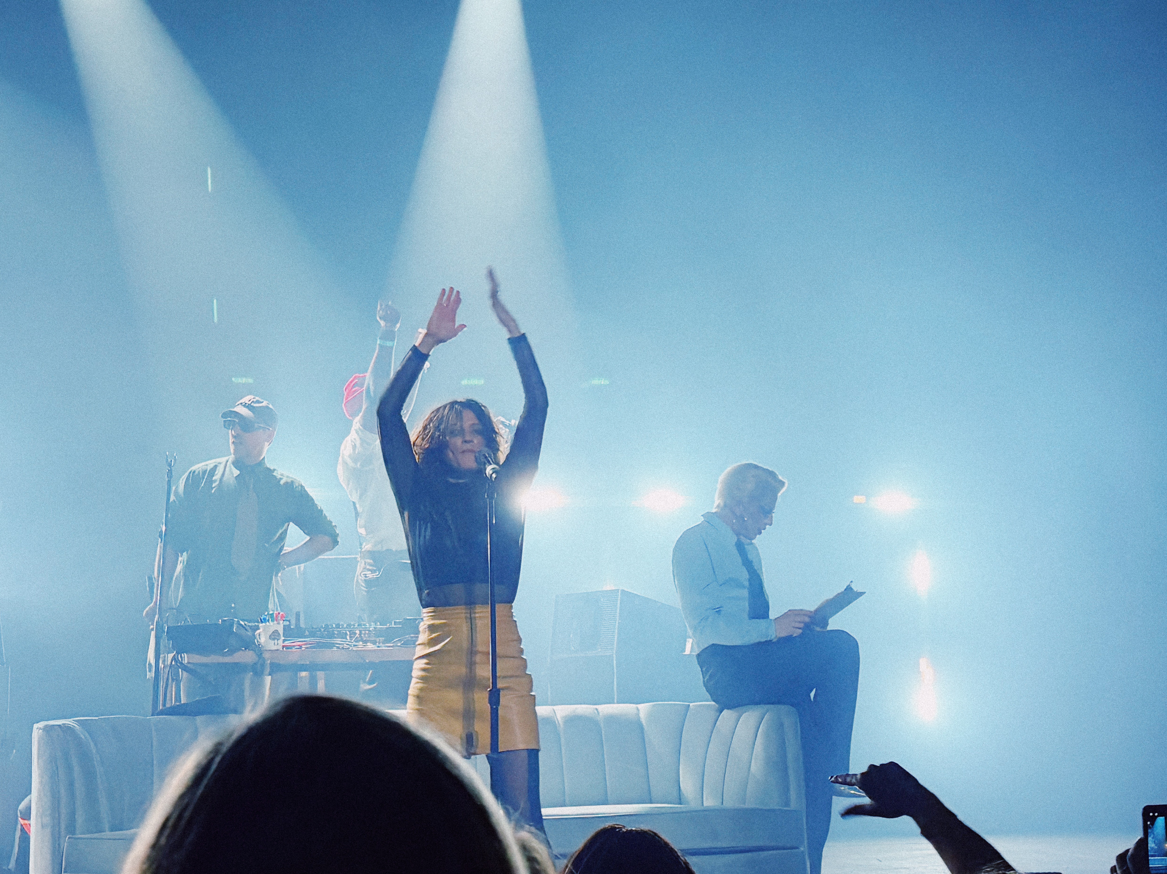 Dragonette stands at a mic at center stage, wearing a yellow skirt and black sheer top, arms raised as she sings. Behind her, two members of The Knocks work at a DJ table, one in a cap and tie, the other wearing a red beanie and raising a fist. To the right, Aquaria with platinum hair sits casually on a white couch reading from a clipboard. The stage is washed in cool blue light with bright beams behind them.