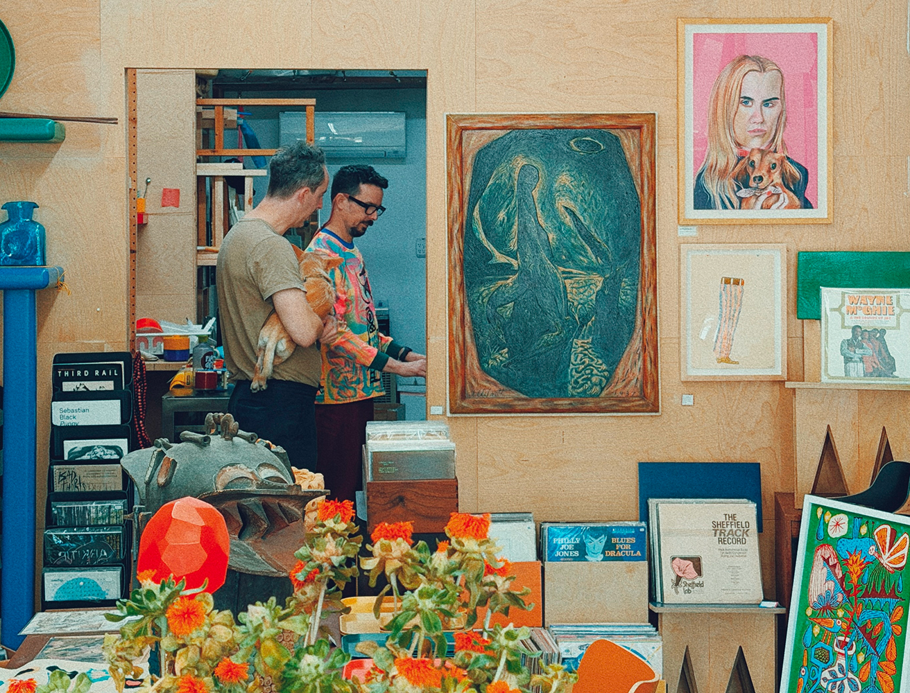 Inside the Face Guts shop, two people stand near the back discussing work while holding a small dog. The space is filled with art on every surface—framed portraits, abstract paintings, records, and sculpture. A table of orange flowers and mismatched chairs sits in the foreground, giving the studio a warm, lived-in creative energy.