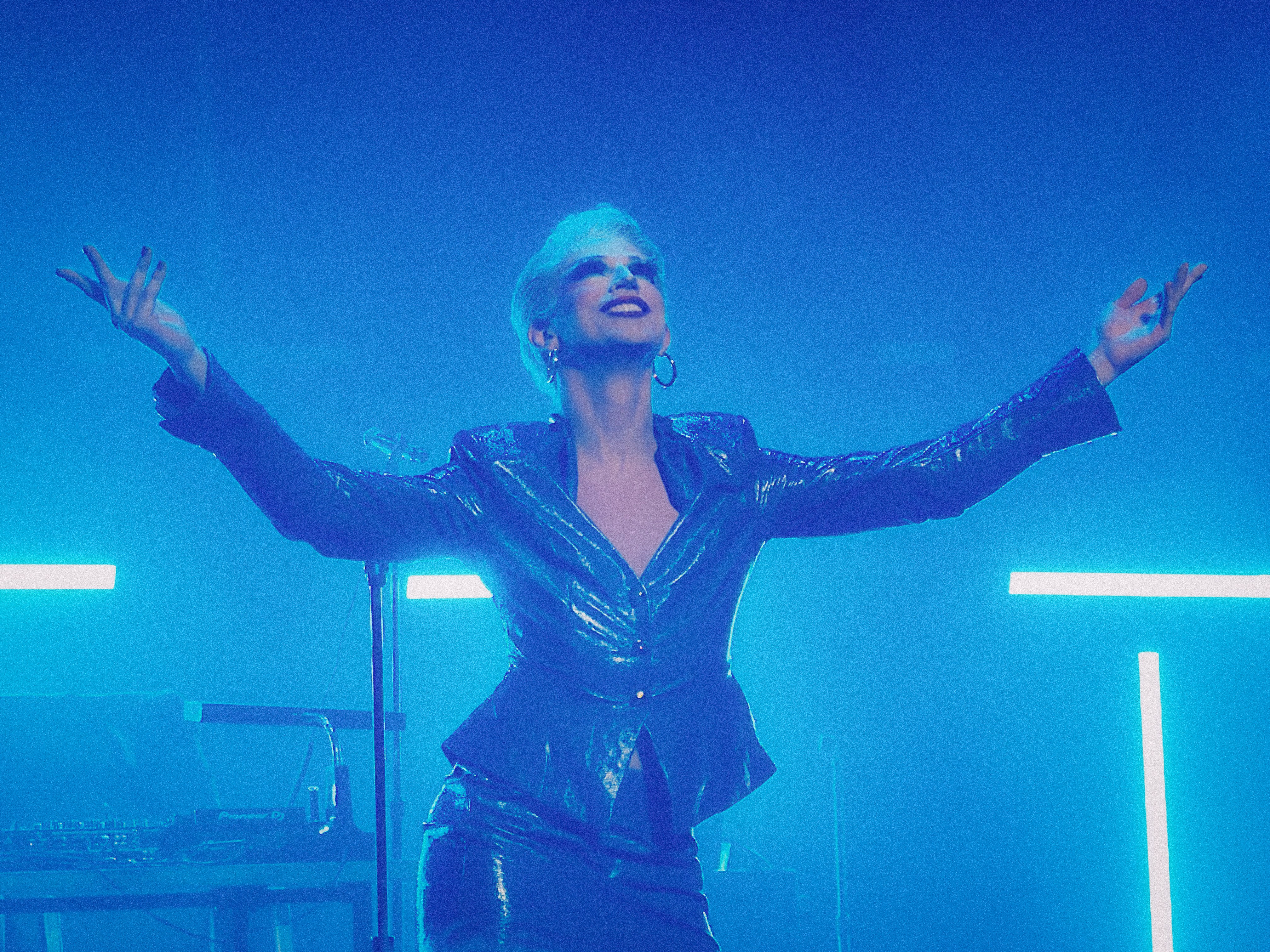 Aquaria poses triumphantly with arms wide open during her Keynote Adress, wearing a shiny, structured blue jacket and matching skirt. Her short platinum hair and bold makeup catch the bright icy blue stage lights, giving her an almost ethereal, futuristic, EXECUTIVE presence.